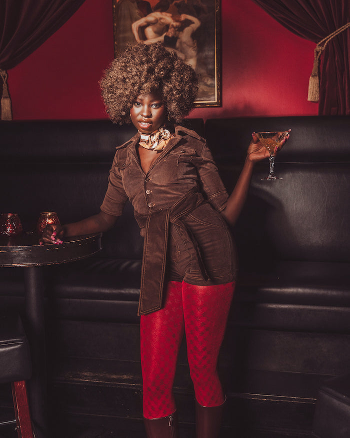 Woman in a brown coat and red pants holding a cocktail in a dimly lit room.