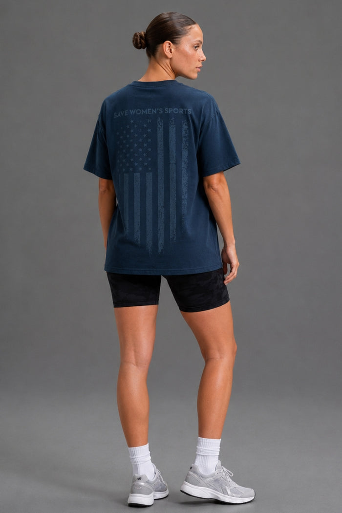 A woman shown from the back wearing a navy blue athletic T-shirt with a faded American flag graphic and 'WOMEN'S SPORTS' text, paired with black shorts, white socks, and gray sneakers against a gray studio background. || Navy