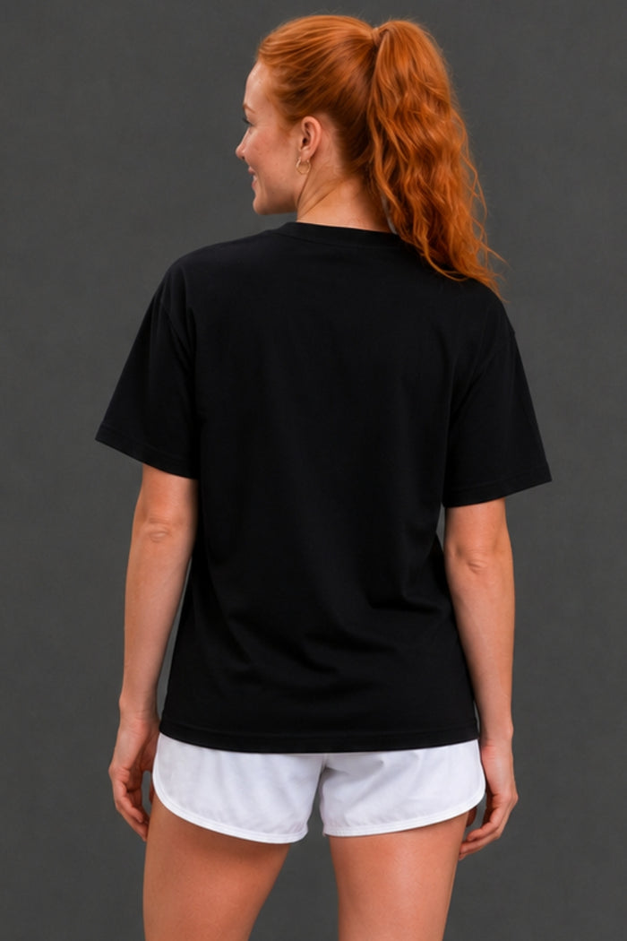 A woman with red hair in a ponytail is shown from the back, wearing a plain black short-sleeve t-shirt and white athletic shorts against a dark gray studio background. || Black