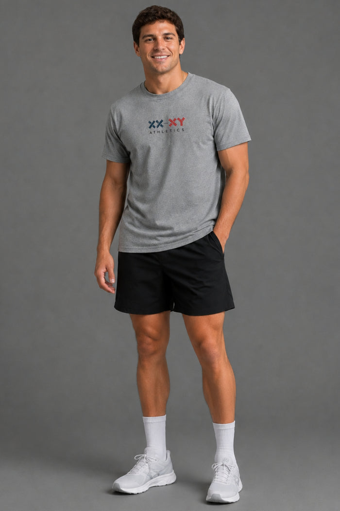 A man modeling a heather gray short-sleeve t-shirt with a 'XX-XY Athletics' logo on the chest, paired with black athletic shorts, white socks, and white sneakers, against a solid gray studio background. || Heather Gray