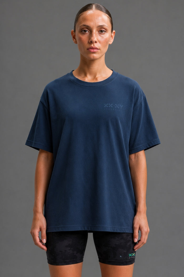 A woman modeling an oversized navy blue t-shirt with a subtle tonal logo on the left chest, paired with black athletic shorts, against a solid gray studio background. || Navy