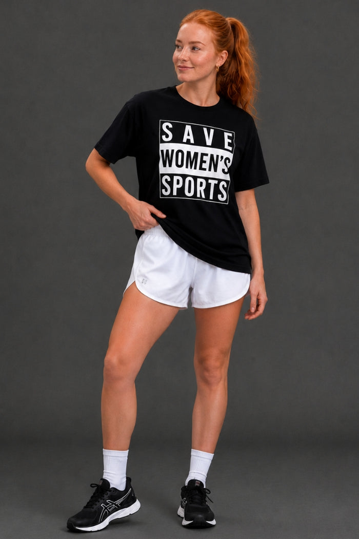 A woman modeling a black graphic t-shirt with white text that reads 'SAVE WOMEN'S SPORTS', paired with white athletic shorts, white socks, and black sneakers, against a dark gray studio background. || Black