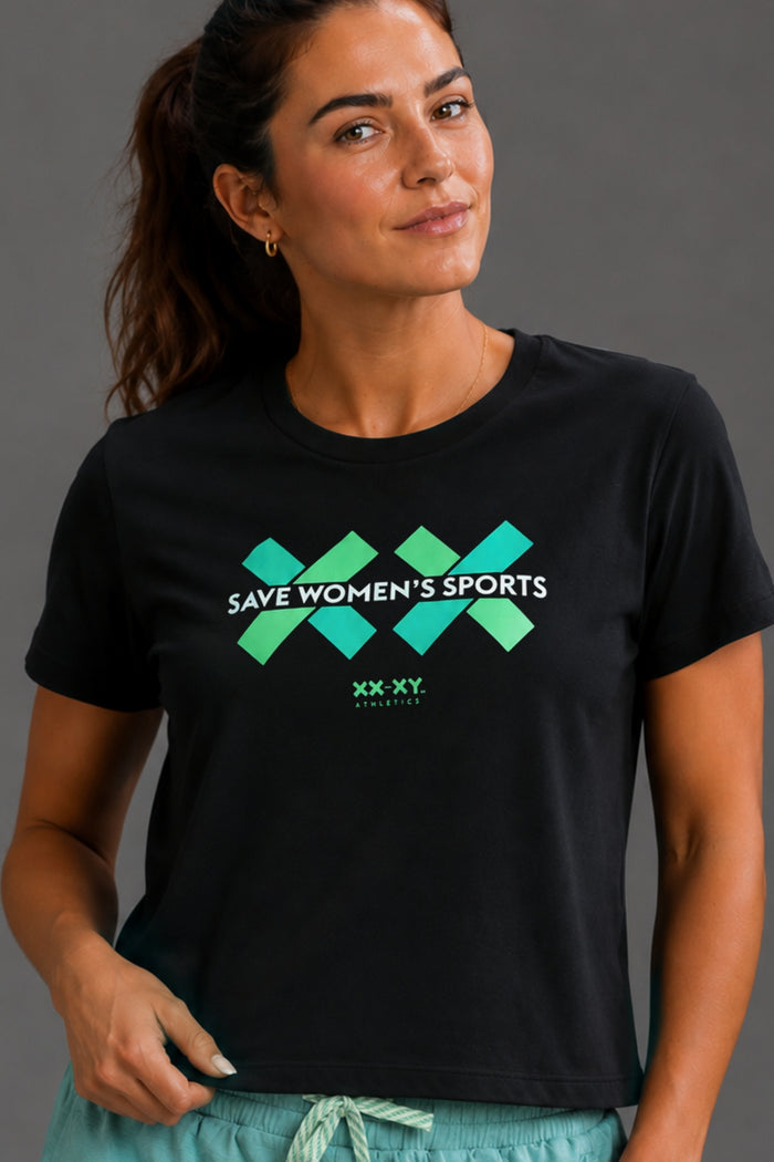 Woman wearing a black shirt with a "SAVE WOMEN'S SPORTS" text and green and blue graphics. Paired with blue shorts on a gray background. || black